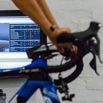 Ciclista rodando sobre el rodillo, en pleno estudio biomecánico. De fondo, se observa un ordenador con el registro de medidas, ángulos y gráficas, obtenidos en tiempo real por 3DMA Cycling.